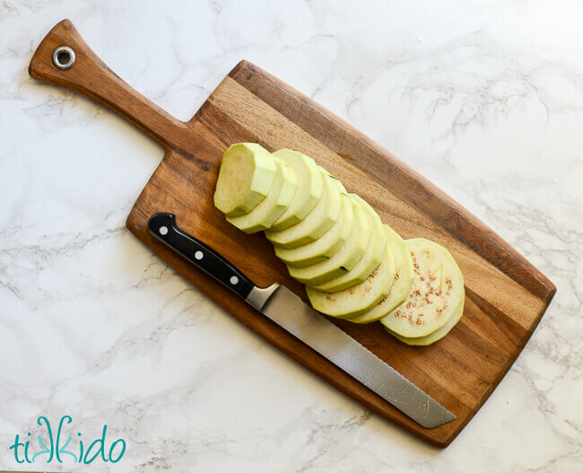 Peeled eggplant, sliced on a wooden cutting board on a white marble surface.