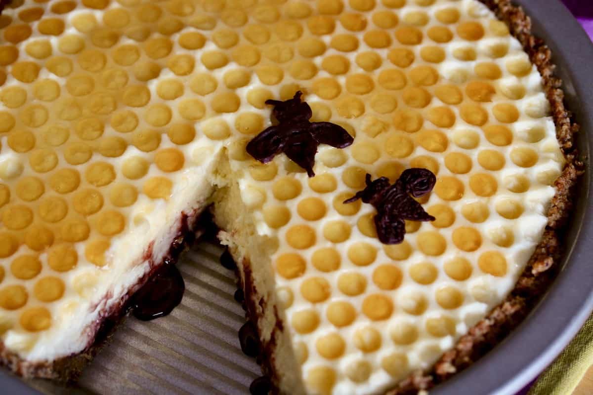 Elderberry honeycomb cream pie in a pie tin with one slice cut out.  The top of the pie looks patterned like a honeycomb with honey drizzled all over the top, and two chocolate bees garnish the pie.
