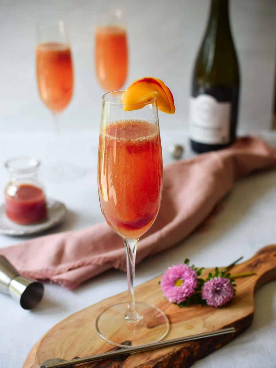 Fresh plum bellini cocktail in a champagne glass, garnished with a slice of plum.  A bottle of champagne, plum puree, and two more glasses of plum bellini are in the background.