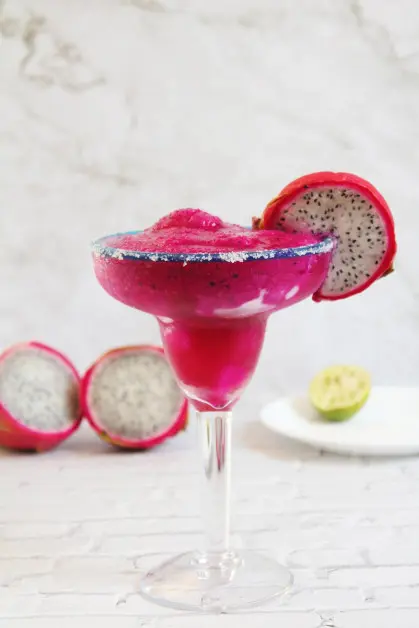 Hot pink frozen dragon fruit margarita in a margarita glass rimmed with salt, and garnished with a slice of dragon fruit. 