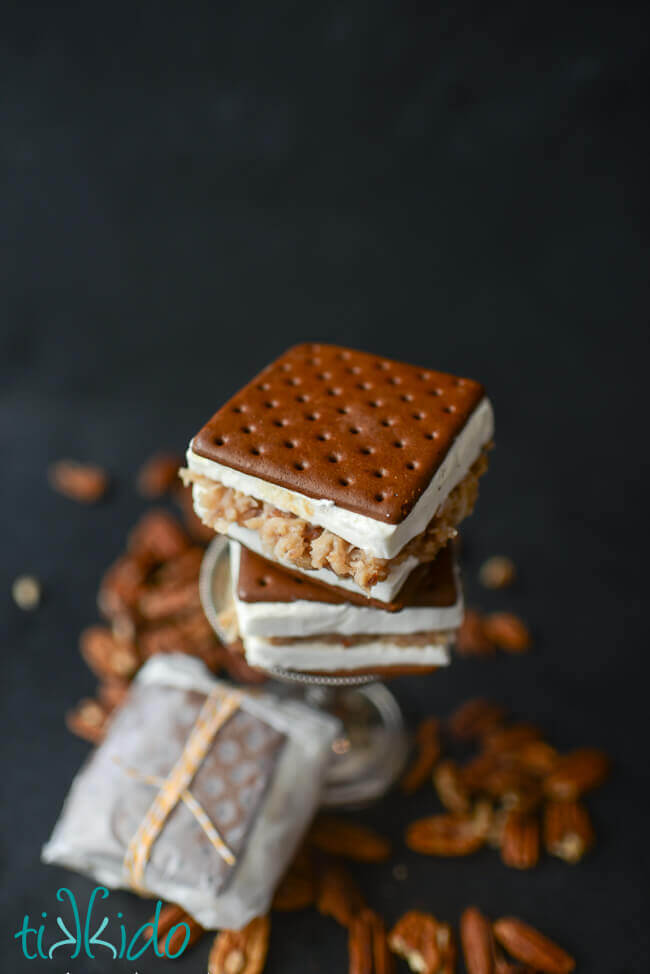 Two ice cream sandwiches filled with German chocolate cake filling stacked on a small metal serving tray, another wax paper wrapped sandwich on the surface blow, pecans scattered around.