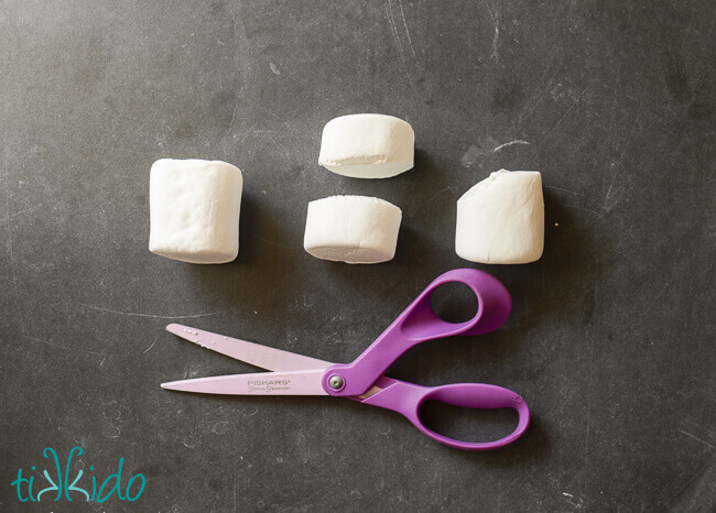Scissors and marshmallows on a black chalkboard surface, showing how to cut the marshmallows to make Ghostbusters Stay Puft Marshmallow Treats.