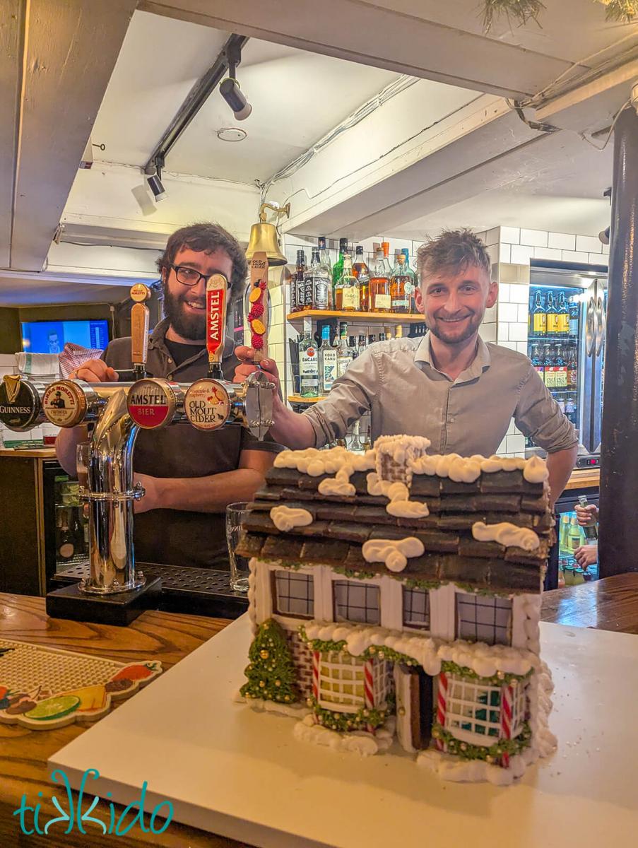 Two happy employees of The Beehive pub in St Albans being surprised with a gingerbread replica of the pub.  The gingerbread house sits on the bar.