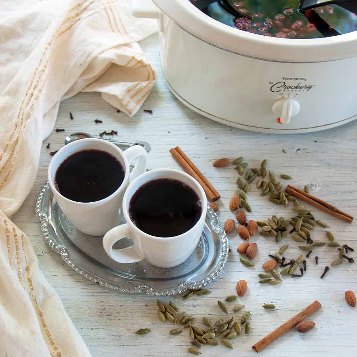 Two mugs filled with slow cooker Scandinavian glogg, surrounded by whole spices, nuts, and a crock pot full of the drink.