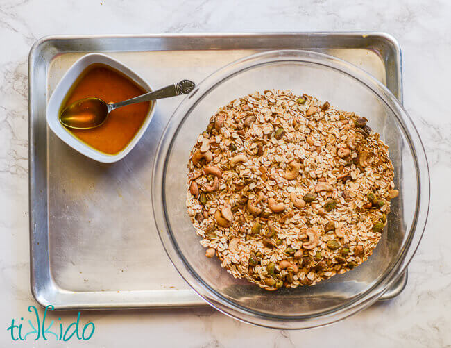 Dry ingredients for Granola Recipe in a bowl, with wet ingredients for the homemade granola combined in a second bowl next to the first.  Both bowls sit in an empty baking sheet.