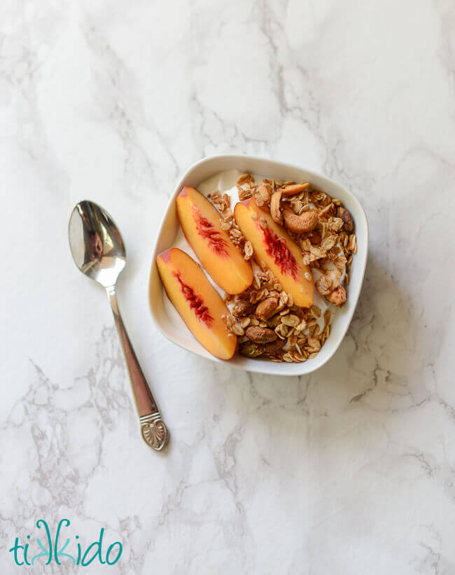 White bowl filled with greek yogurt, homemade granola, and three slices of peach, sitting on a white marble surface next to a silver spoon.