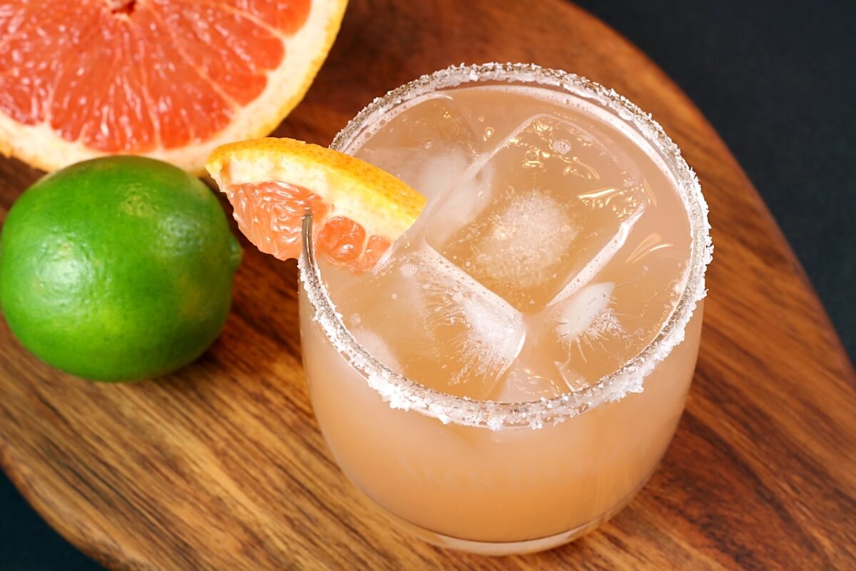 Salt-rimmed glass filled with ice and fresh grapefruit margarita, garnished with a wedge of pink grapefruit, sitting on a wooden cutting board.