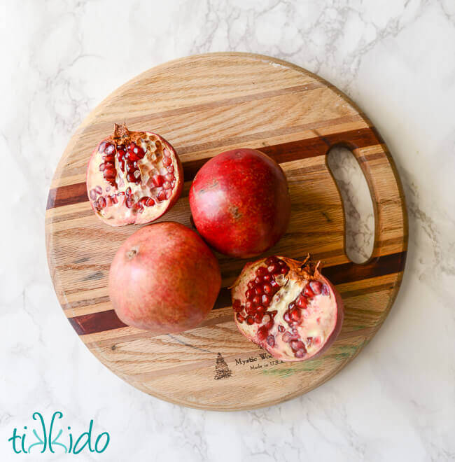 Three pomegranates, two whole, one cut in half, on a wooden cutting board.