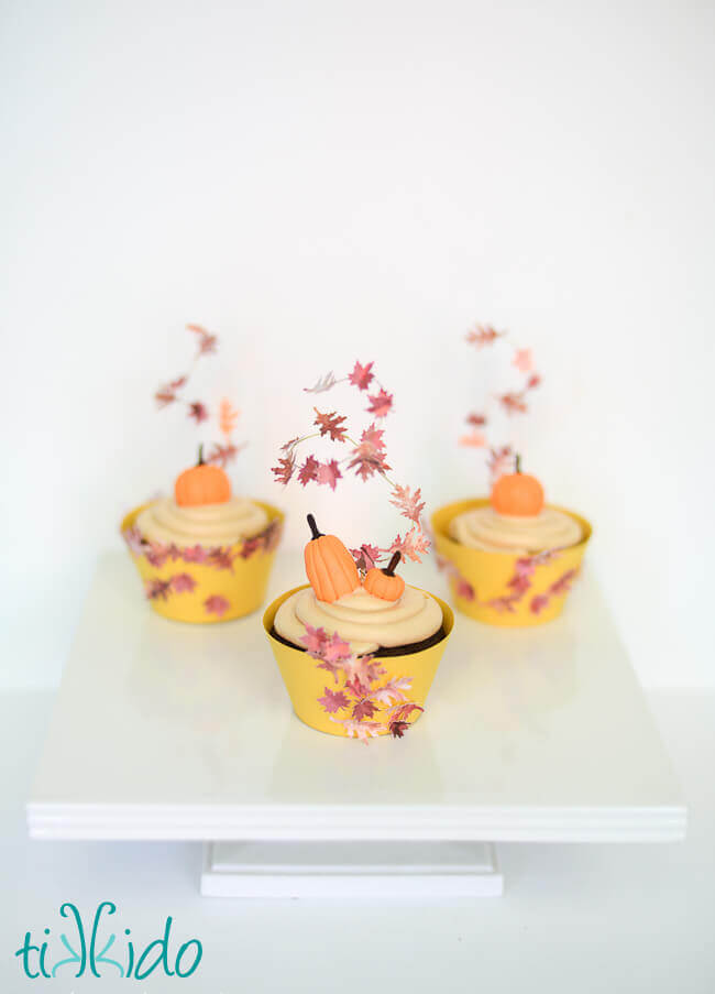 Three cupcakes decorated with Gum Paste Pumpkins and swirling fall leaves cupcake toppers.