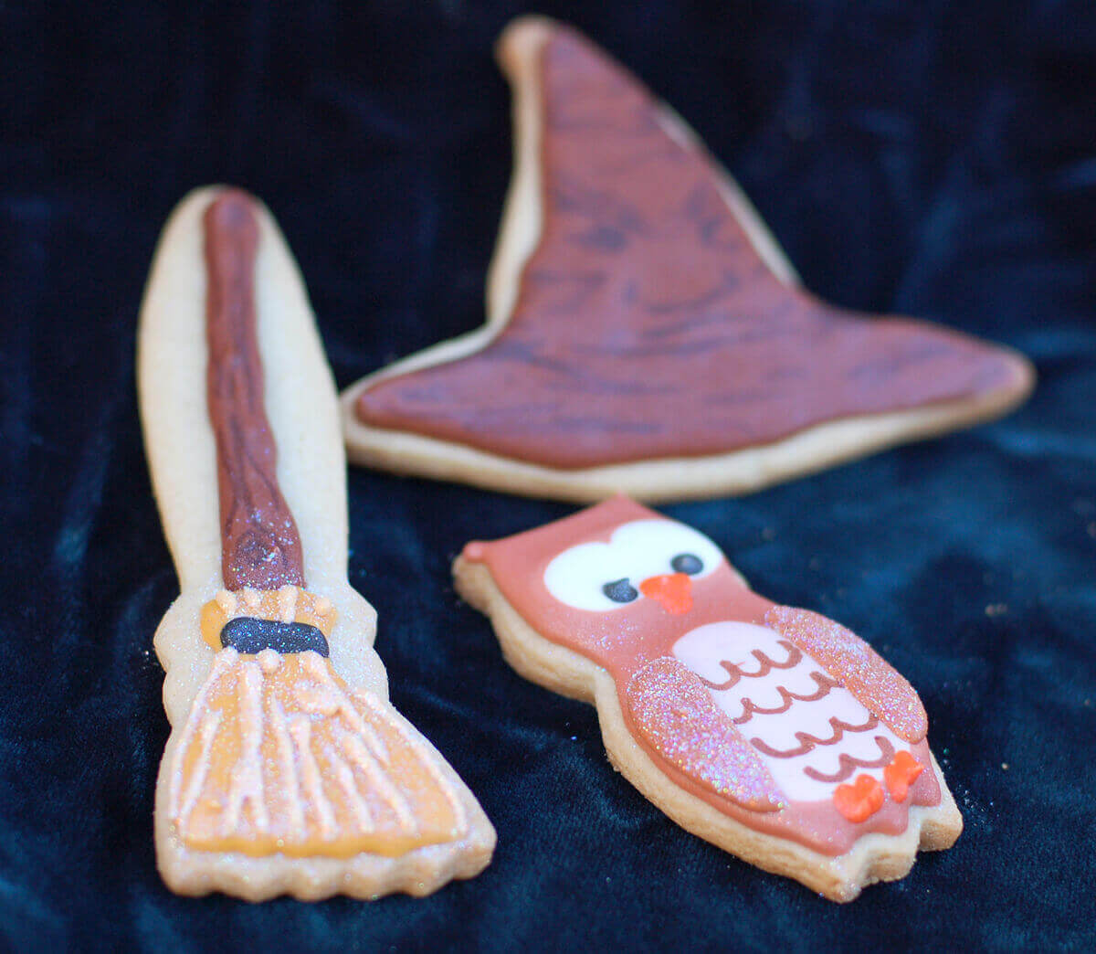 Owl, broom, and sorting hat harry potter sugar cookies on a dark blue velvet background.