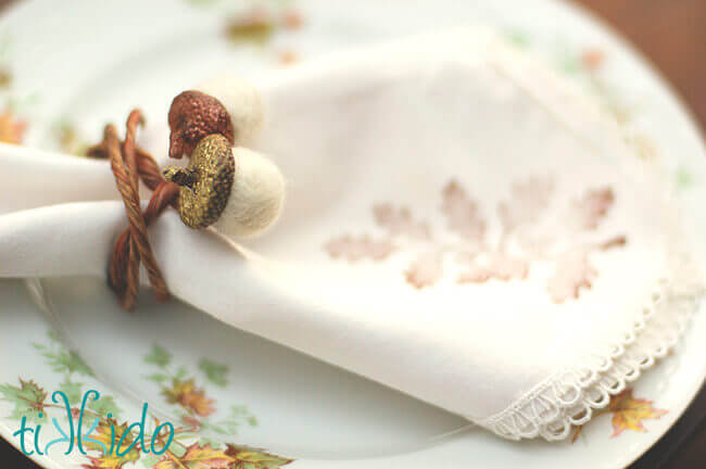 Felt acorn napkin ring for a Thanksgiving dinner on a fabric napkin, on a plate decorated with fall leaves.