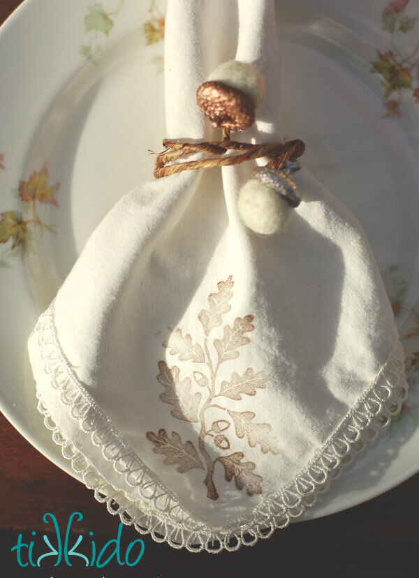 DIY Felt Acorn Thanksgiving Napkin Ring on a napkin stamped with an oak leaf pattern, on a china plate decorated with leaves.