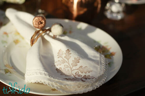 DIY Felt Acorn Thanksgiving Napkin Ring on a napkin stamped with an oak leaf pattern, on a china plate decorated with leaves.