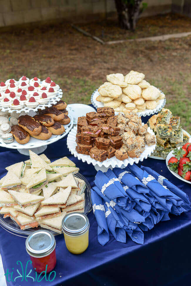 Tea sandwiches, scones, and quiche on white cake stands on a table covered in a blue tablecloth