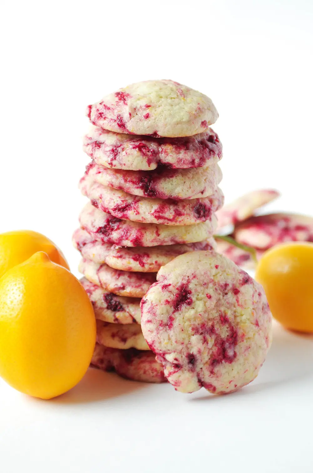 Stack of lemon raspberry cookies surrounded by whole lemons on a white background.