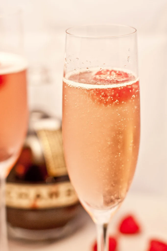 Glass of a light pink French Kiss Cocktail in a champagne glass, with a raspberry floating on top.