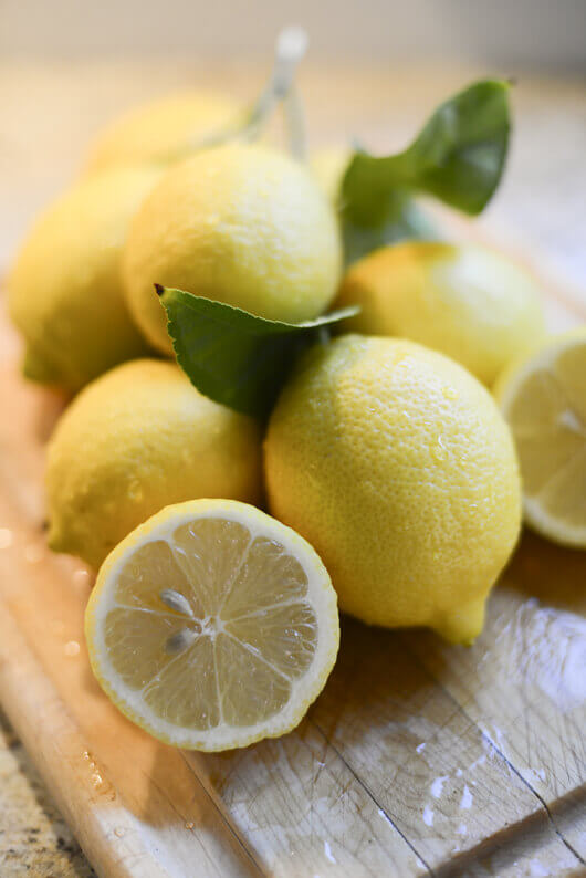 Pile of fresh lemons, whole and cut in half, on a wooden cutting board.