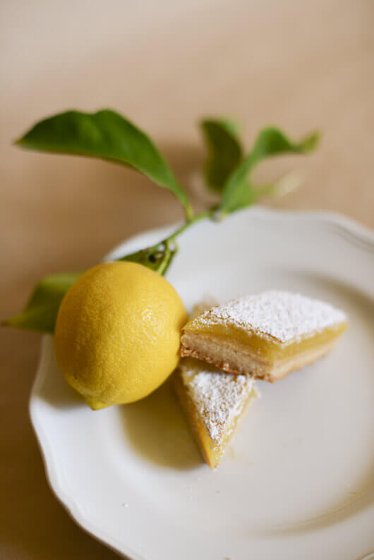 Two lemon bars cut into diamond shapes on a white plate next to a fresh lemon with leaves attached to the stem.