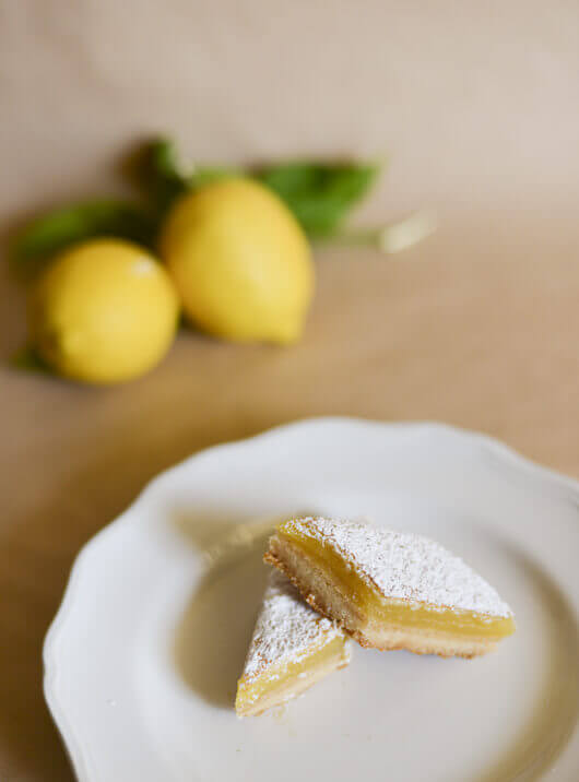 Two lemon bars on a white plate, with two fresh lemons in the background.