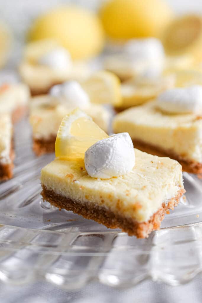 Lemon cheesecake bars on a glass cake plate, topped with whipped cream and a slice of lemon.