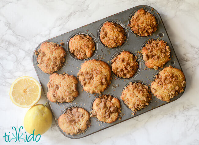 Muffin tin with lemon muffins with crumb topping on a white marble background next to fresh lemons.