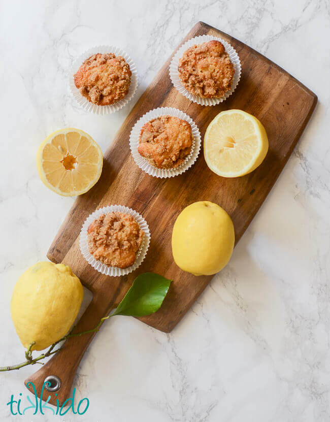 Fresh lemons and lemon muffins on a wooden cutting board on a white marble surface.