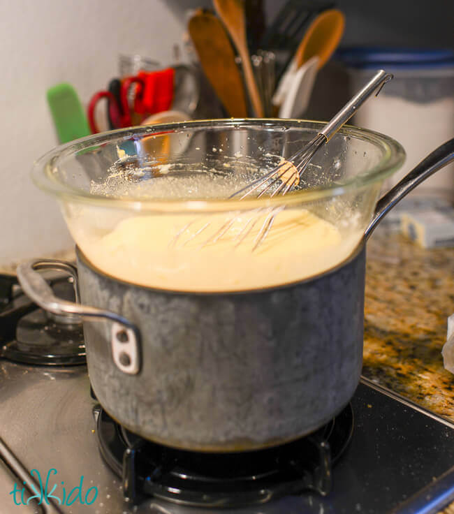 Improvised double boiler made with a saucepan and clear pyrex bowl, with lemon curd and whisk in the bowl on a gas stove.