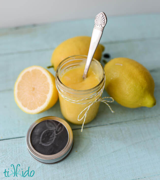 Mason jar with lemon curd and a spoon, beside one lemon cut in half and two full lemons on a light blue wooden background.
