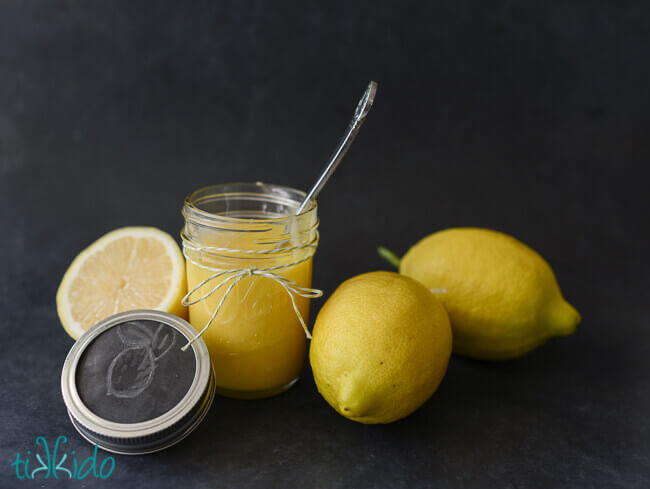 Mason jar of lemon curd with the lid off and a spoon in the har, two lemons and a cut lemon beside it, on a black chalkboard background.