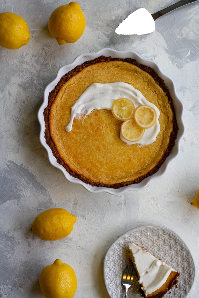 whole lemon pie with graham cracker crust baked in a white pie plate.  Two lemons and a pie server sit next to the pie.