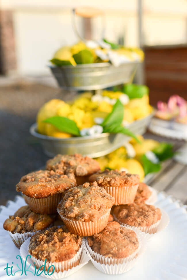 Stack of lemon muffins with crumb topping on a white cake stand in front of a tiered tray full of lemons