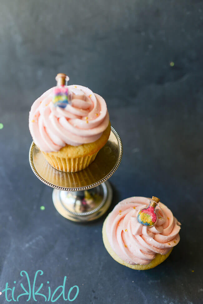 Two vanilla cupcakes topped with light pink icing and a tiny bottle full of multi colored sprinkles.