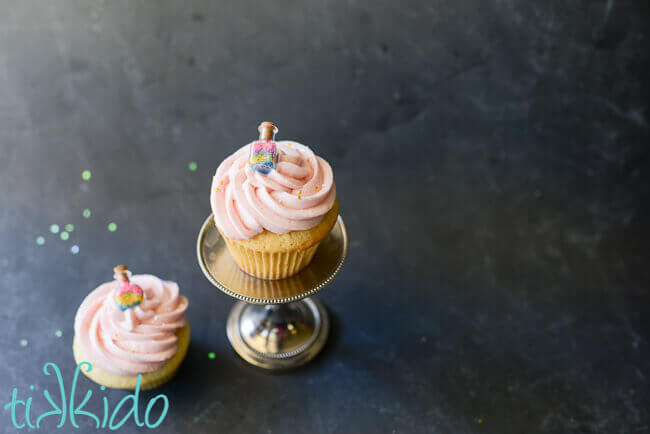 Two vanilla cupcakes topped with light pink icing and a tiny bottle full of multi colored sprinkles.