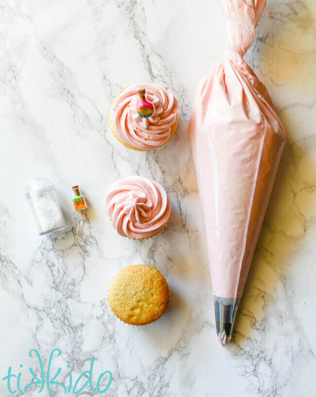 Bag of pink icing with a large star tip, next to three vanilla cupcakes, to of which have pink icing and one of which has a sprinkle potion bottle topper.