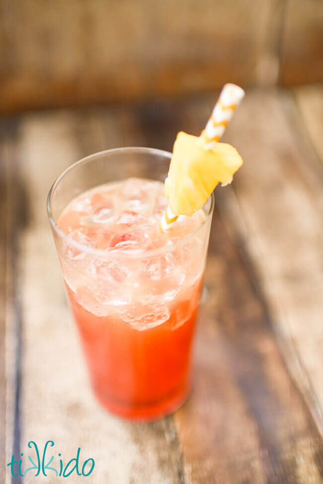 Top view of a summer cocktail in a tall glass made with Malibu coconut rum, pineapple juice, and grenadine syrup, with a yellow and white chevron striped paper straw and a chunk of fresh pineapple.