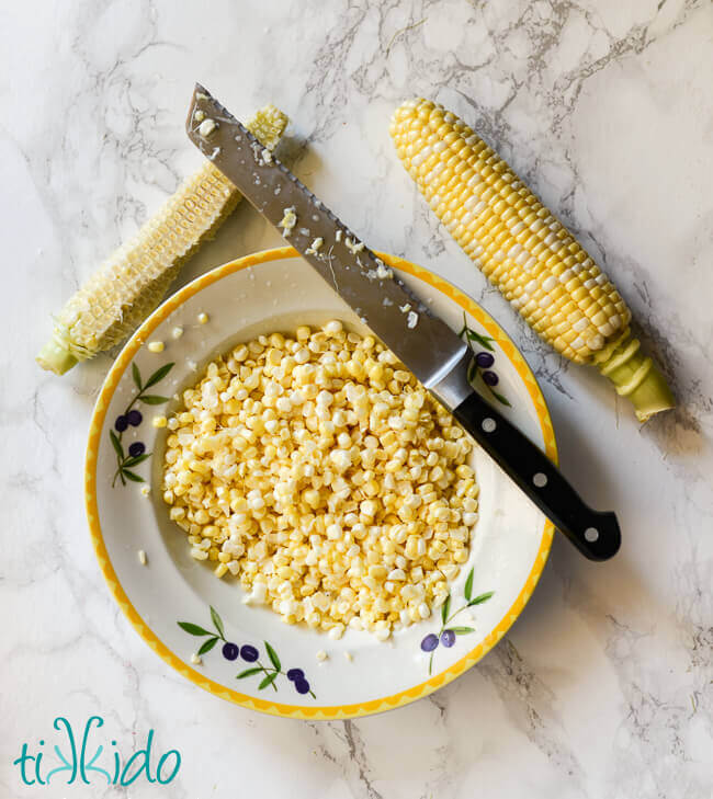 Bowl of sweet corn kernels with a knife balanced on the lip, next to two ears of corn, one of which with the kernels removed.