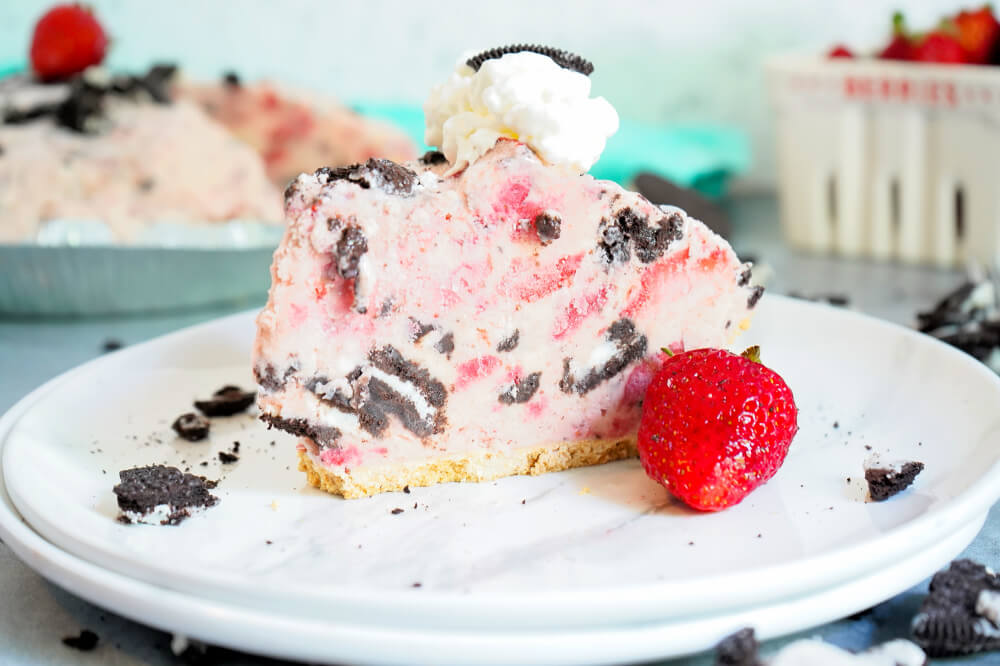 Strawberry Cookies and Cream frozen pie slice on a white plate, garnished with a fresh strawberry, whipped cream, and a chunk of oreo cookie.