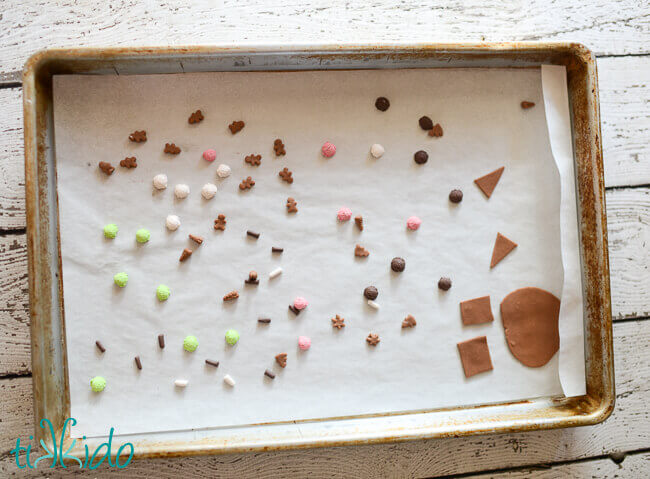 Polymer clay shaped into tiny gingerbread houses and gingerbread cookies, on a lined baking sheet.