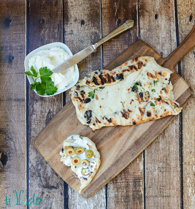 Grilled bread, torn, on a wooden cutting board.  A small piece of the grilled bread is spread with whipped feta spread, and topped with a few green olives.  A bowl of whipped feta spread sits next to the cutting board.