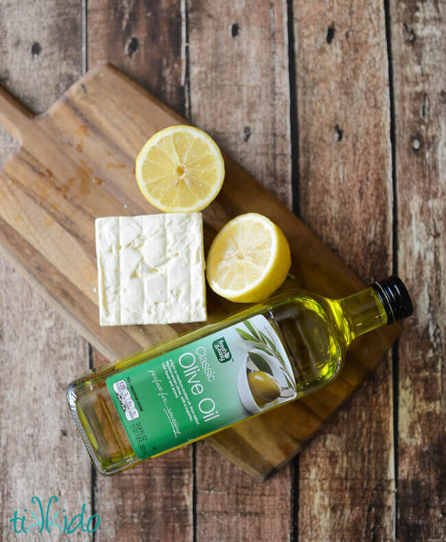 Feta cheese, a sliced lemon, and bottle of olive oil to make whipped feta spread on a wooden cutting board on a wooden table.