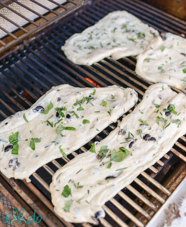 Bread dough covered in olive oil, sea salt, and fresh herbs, being cooked on a grill.