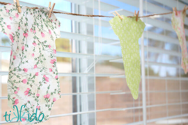 Baby shower garland made from fabric onesie cutouts and miniature clothespin hanging in front of a window.