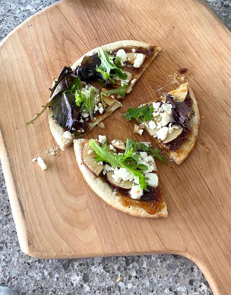 Fig, apple, arugula, and goat cheese flat bread cut into appetizer sized slices on a wooden cutting board.