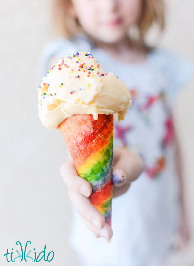 Girl holding an ice cream cone painted with rainbow stripes, filled with vanilla ice cream and topped with rainbow sprinkles.