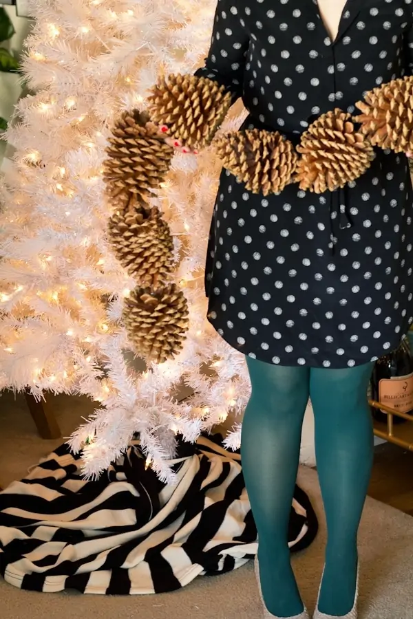 woman in a black and white polka dot dress holding a Christmas garland made from giant pine cones.  She stands in front of a white artificial Christmas tree.