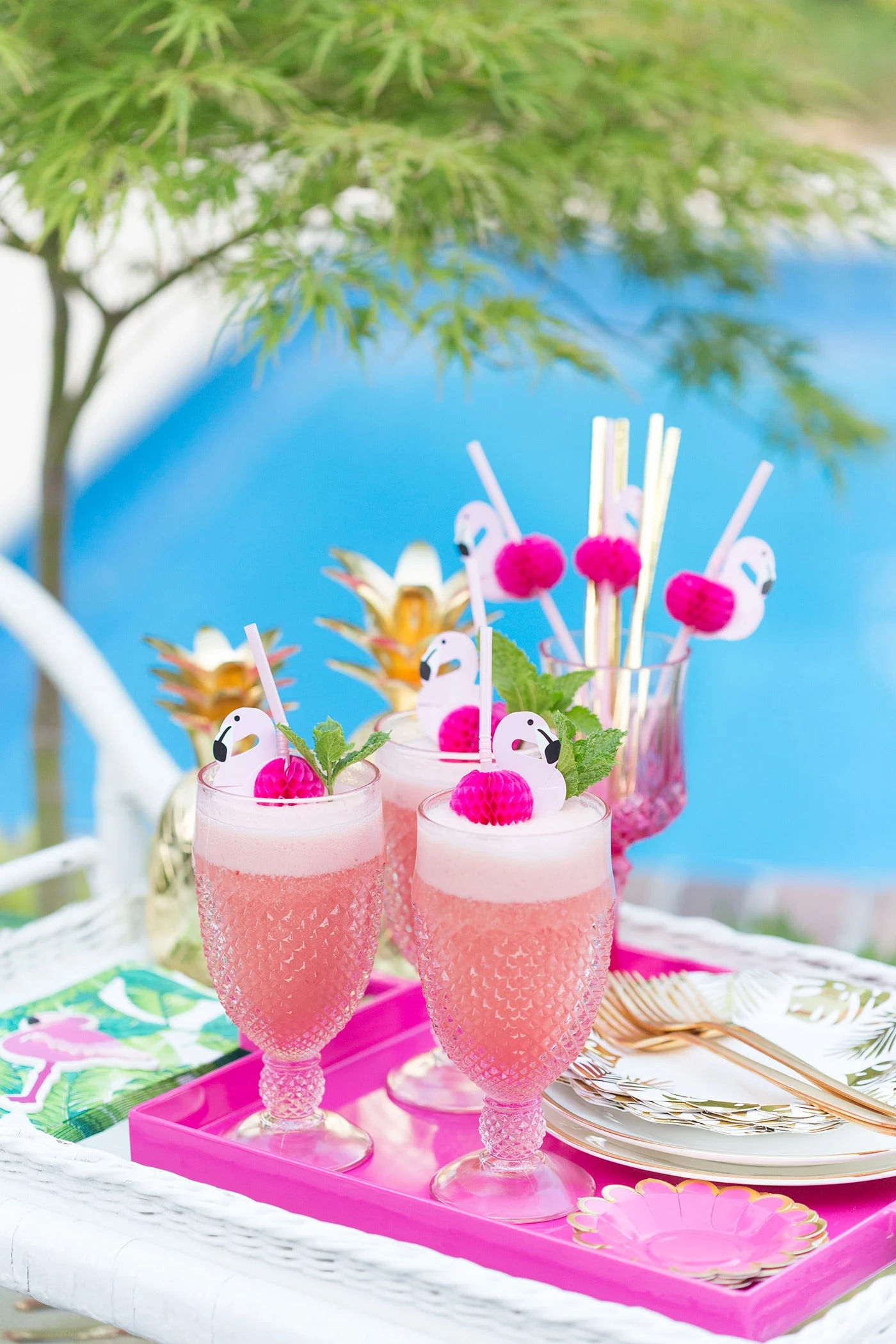 Pink tray with three glasses filled with bright pink Flamingo Punch cocktails, each glass garnished with a straw with a paper flamingo on it.