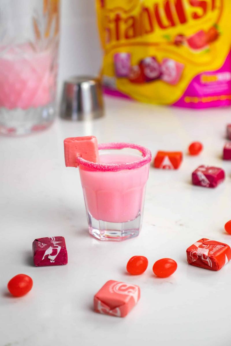 A pink starburst shot in a clear shot glass rimmed with pink sugar, and garnished with a pink Starburst candy.  Other wrapped starburst candies are scattered around the shot glass.