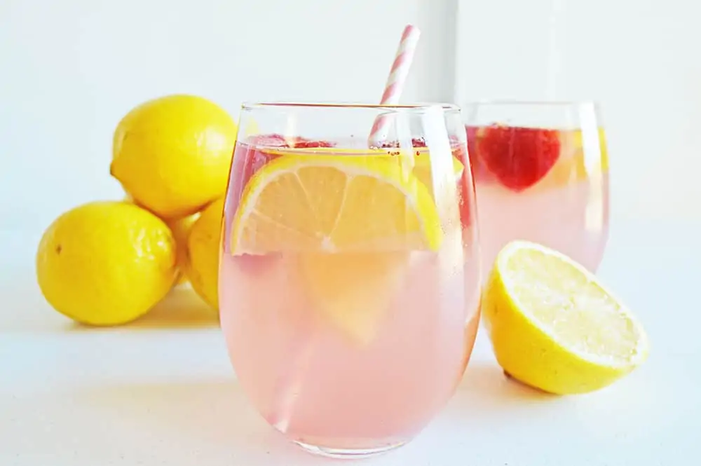 Pink lemonade moscato cocktail in a glass with slices of lemon and fresh raspberries floating in the drink.  Fresh whole lemons are in the background.