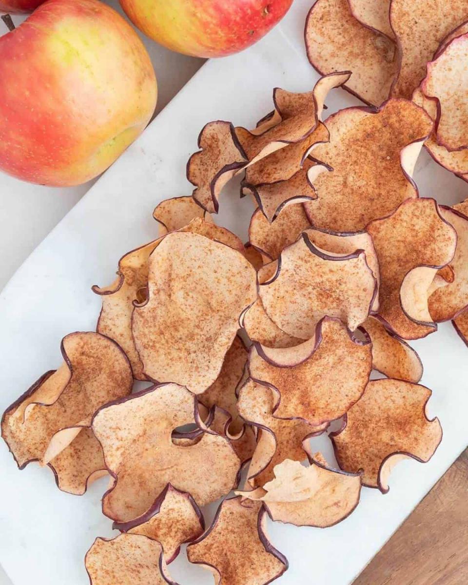 Crispy apple chips dusted with cinnamon on a white plate, next to fresh apples.