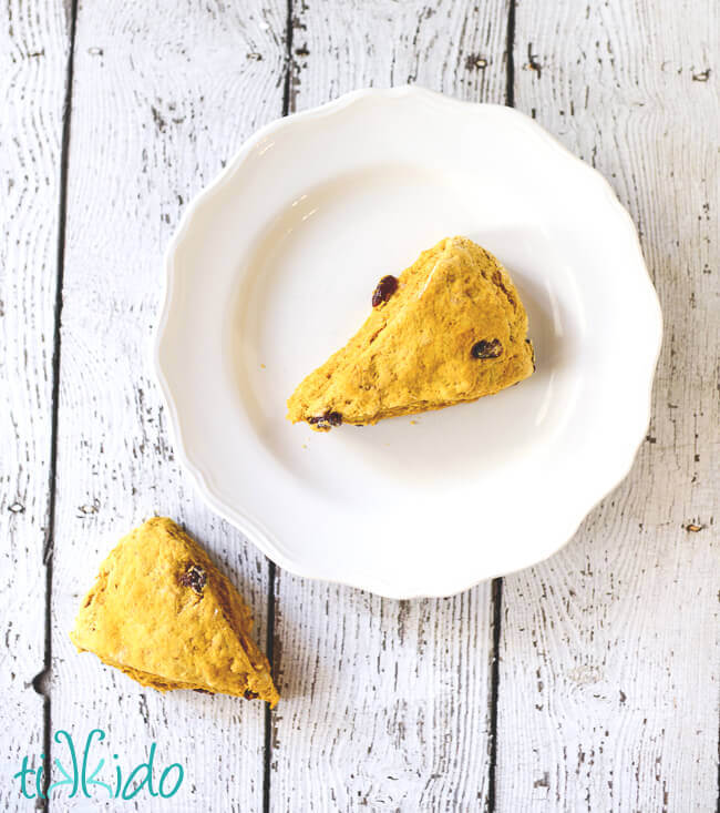 One Pumpkin cranberry scone on a white plate, a second on the white weathered wood table beside the plate.